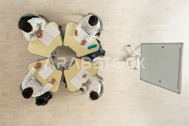 تأدية الواجبات المدرسية، بورتريه علوي عامودي لتلاميذ عرب خليجيون سعوديون يرتدون الثوب السعودي التقليدي  يجلسون على الكراسي وأمامهم طاولات الدراسة، الاهتمام بالعلم والتعلم، شغف التعاون والمساعدة، خلفية بيج