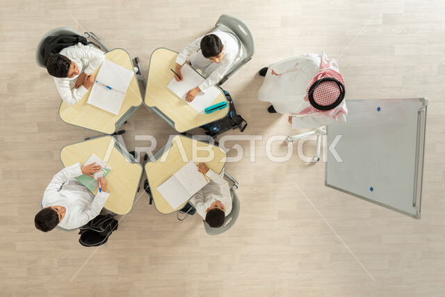 تأدية الواجبات المدرسية، تلاميذ عرب خليجيون سعوديون يرتدون الزي السعودي التقليدي، الانتباه الى المدرس، بورتريه علوي عامودي لمعلم عربي خليجي سعودي يقوم بمساعدة الطلاب بعمل نشاطات جماعية، الاهتمام بالعلم والتعلم، مفهوم الاجتهاد و طلب التفوق، خلفية بيج