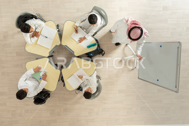 الاهتمام بالعلم والتعلم، مفهوم الاجتهاد و طلب التفوق، تلاميذ عرب خليجيون سعوديون يرتدون الزي السعودي التقليدي، شدة الانتباه الى الدرس، بورتريه علوي عامودي لمعلم عربي خليجي سعودي يقوم بمساعدة الطلاب بعمل نشاطات جماعية، خلفية بيج