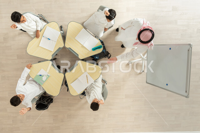 مفهوم الاجتهاد و طلب التفوق، تنمية المهارات الابداعية، تلاميذ عرب خليجيون سعوديون يرتدون الزي السعودي التقليدي يرفعون أيديهم للمشاركة، القيام بكتابة الواجبات المدرسية، بورتريه علوي عامودي لمعلم عربي خليجي سعودي يقوم بمساعدة الطلاب بعمل نشاطات جماعية، خلفية بيج