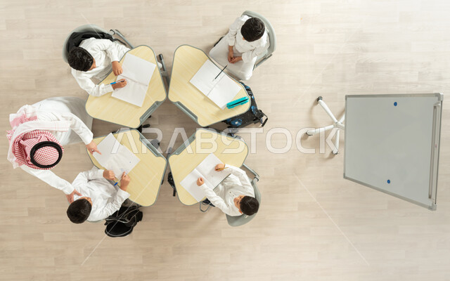 تنمية المهارات الابداعية، تلاميذ عرب خليجيون سعوديون يرتدون الزي السعودي التقليدي، القيام بكتابة الواجبات المدرسية، بورتريه علوي عامودي لمعلم عربي خليجي سعودي يقوم بمساعدة الطلاب بعمل نشاطات جماعية، مفهوم الاهتمام بالعلم والتعليم، خلفية بيج