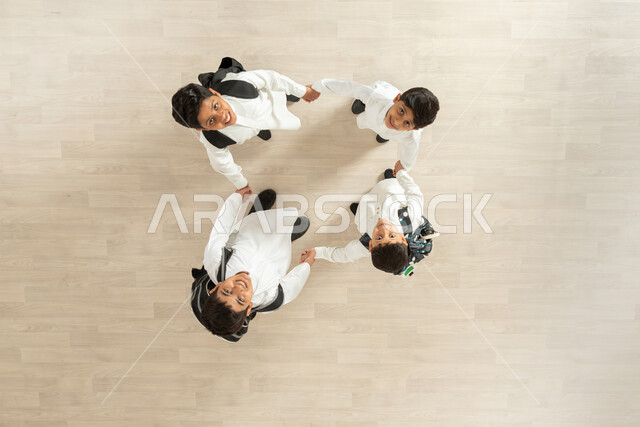 النظر الى الكاميرا بوجوه تعلوها الابتسامة وايماءات الفرح والسعادة، بورتريه علوي عامودي لمجموعة أولاد عربيين خليجيين سعوديين يرتدون الثوب التقليدي يقفون متلاصقين ويمسكون أيدي بعضهم البعض بإحكام، خلفية بيج