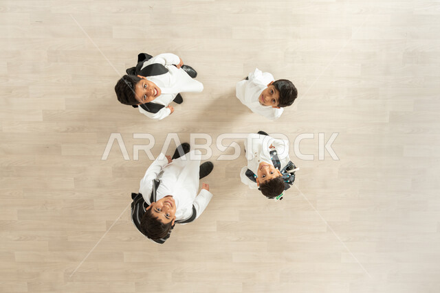 النظر الى الكاميرا بإيماءات السعادة والفرح والسرور، بورتريه علوي عامودي لمجموعة أولاد عربيين خليجيين سعوديين يرتدون الثوب التقليدي و يحملون على ظهرهم الحقيبة المدرسية، خلفية بيج