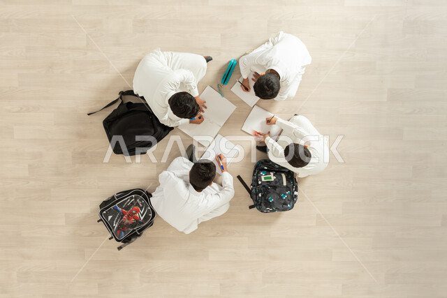 Writing homework, developing creative skills, a vertical portrait of a group of Saudi Gulf Arab boys wearing traditional thobes sitting on the floor, enjoying group recreational activities, beige background