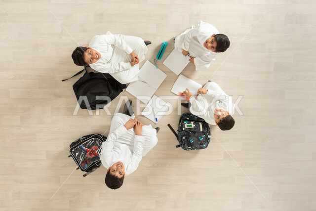 الاستمتاع  بالأنشطة الترفيهية الجماعية, بورتريه علوي عامودي لمجموعة أولاد عربيين خليجيين سعوديين يرتدون الثوب التقليدي  يجلسون على الأرض, النظر باتجاه الكاميرا بتعابير وجوه مبتسمة مليئة بالسرور, عرض شاشة سوداء فارغة عبر الحاسوب و الجهاز اللوحي, خلفية بيج