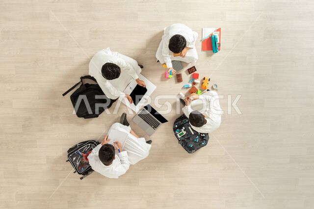 Using modern devices in studying, sharing with friends in learning and playing, a vertical overhead portrait of a group of Saudi Gulf Arab boys wearing traditional thobes sitting on the floor, displaying a blank black screen on the computer and tablet, beige background