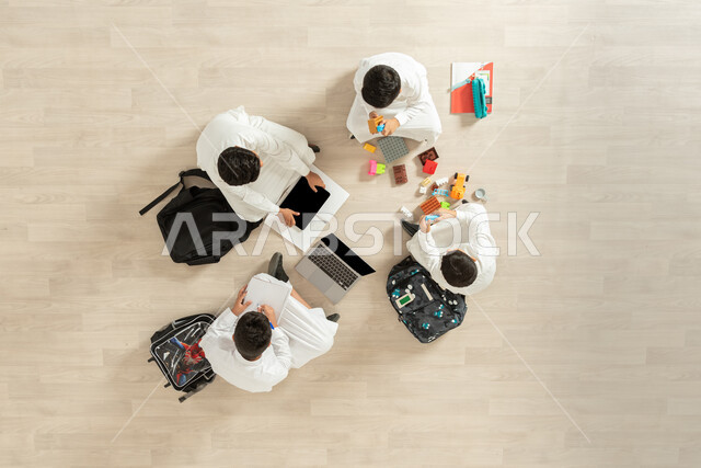 مشاركة الأصدقاء في التعلم و اللعب, استخدام الأجهزة الحديثة في الدراسة, بورتريه علوي عامودي لمجموعة أولاد عربيين خليجيين سعوديين يرتدون الثوب التقليدي  يجلسون على الأرض, عرض شاشة سوداء فارغة عبر الحاسوب و الجهاز اللوحي, خلفية بيج