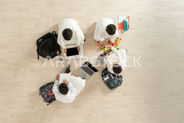 مشاركة الأصدقاء في التعلم و اللعب , استخدام الأجهزة الحديثة في الدراسة , بورتريه علوي عامودي لمجموعة أولاد عربيين خليجيين سعوديين يرتدون الثوب التقليدي  يجلسون على الأرض , عرض شاشة سوداء فارغة عبر الحاسوب و الجهاز اللوحي , خلفية بيج