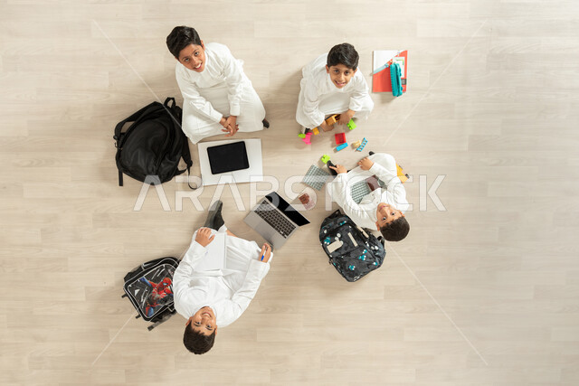 الاستمتاع  بالأنشطة الترفيهية الجماعية , بورتريه علوي عامودي لمجموعة أولاد عربيين خليجيين سعوديين يرتدون الثوب التقليدي  يجلسون على الأرض , النظر إلى الكاميرا بتعابير السعادة و السرور , عرض شاشة سوداء فارغة عبر الحاسوب و التابلت , خلفية بيج
