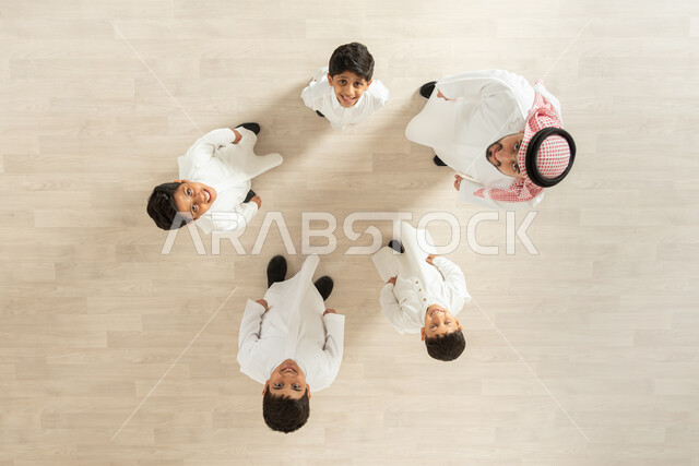 النظر إلى الكاميرا بإيماءات السعادة و السرور , أجواء عائلية سعيدة , بورتريه علوي عامودي لأب عربي خليجي سعودي يرتدي الثوب التقليدي و الشماغ يقوم باللعب مع أبنائه , ممارسة الألعاب الجماعية الممتعة , تعزيز الروابط الأسرية , خلفية بيج