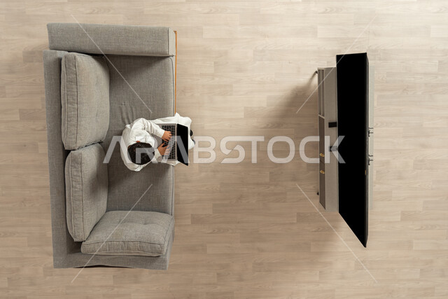 Completing lessons and school assignments using technical devices, a vertical portrait of a Saudi Gulf Arab boy wearing a traditional dress sitting on the sofa in the living room holding a computer in his hands, distance learning, displaying a blank black screen on the TV and computer, beige background