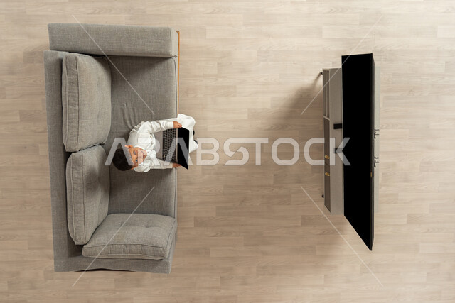 التعلم عن بعد عبر الأجهزة التقنية الحديثة , بورتريه علوي عامودي لولد عربي خليجي سعودي يرتدي الثوب التقليدي  يجلس على الأريكة في غرفة المعيشة يمسك جهاز الحاسوب بيديه  ينظر باتجاه الكاميرا بتعابير وجه مبتسم  , عرض شاشة سوداء فارغة عبر التلفاز و الحاسب , خلفية بيج