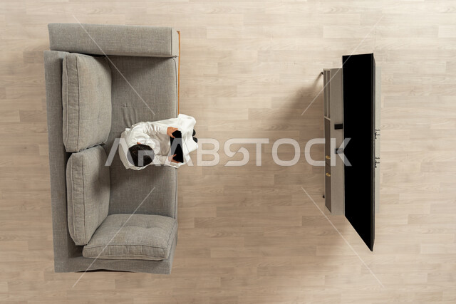 استخدام التكنولوجيا للتسلية و تعبئة الوقت , بورتريه علوي عامودي لولد عربي خليجي سعودي يرتدي الثوب التقليدي  يجلس على الأريكة في غرفة المعيشة يمسك الجهاز اللوحي بيديه  , لعب ألعاب الفيديو المسلية و الممتعة , عرض شاشة سوداء فارغة عبر التلفاز و التابلت , خلفية بيج