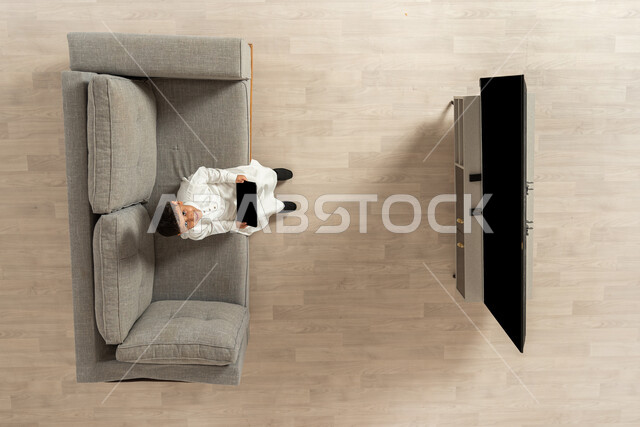 الترفيه و اللعب باستخدام التكنولوجيا الحديثة , ألعاب الفيديو المسلية , بورتريه علوي عامودي لولد عربي خليجي سعودي يرتدي الثوب التقليدي يجلس على الأريكة في غرفة المعيشة يمسك الجهاز اللوحي بيديه و ينظر إلى الكاميرا بإيماءات السعادة , عرض شاشة سوداء فارغة عبر التلفاز و التابلت , خلفية بيج