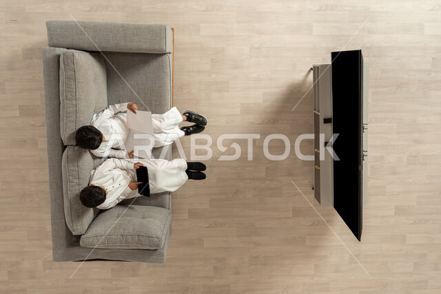 مشاركة الأصدقاء في هواية المطالعة , القيام بالأنشطة الترفيهية المنزلية , بورتريه علوي عامودي لولدان عربيان خليجييان سعوديان يرتديان الثوب التقليدي يجلسان على الأريكة في غرفة المعيشة , تنمية مهارات القراءة عند الأولاد عن طريق الكتب و التقنيات الحديثة ,عرض شاشة سوداء فارغة عبر التلفاز و الجهاز اللوحي , خلفية بيج