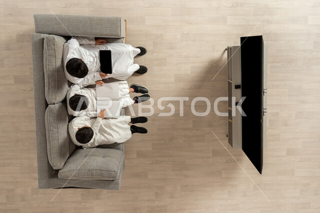 Learning using modern technology, practicing the hobby of reading through a tablet and books, a vertical overhead portrait of three Saudi Gulf Arab boys wearing traditional thobes sitting on the sofa in the living room reading entertaining stories, pointing by hand, integrating and sharing, showing a blank black screen on the TV And the tablet, beige background