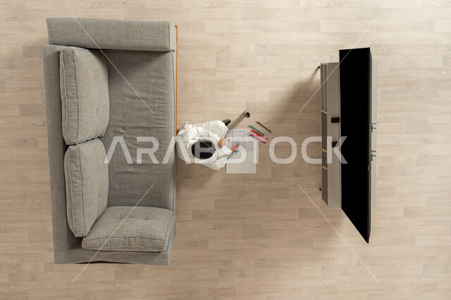 ممارسة هواية الرسم و التلوين في أوقات الفراغ , بورتريه علوي عامودي لولد عربي خليجي سعودي يرتدي الثوب التقليدي  يجلس على الأرض في غرفة المعيشة يقوم بالرسم على الدفتر, القيام بالأنشطة الترفيهية الممتعة و تنمية المواهب , عرض شاشة سوداء فارغة عبر التلفاز , خلفية بيج