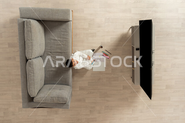 ممارسة الأنشطة الترفيهية المنزلية , بورتريه علوي عامودي لولد عربي خليجي سعودي يرتدي الثوب التقليدي  يجلس على الأرض في غرفة المعيشة ينظر إلى الكاميرا بإيماءات السعادة و السرور , تنمية هواية الرسم و التلوين في أوقات الفراغ , عرض شاشة سوداء فارغة عبر التلفاز , خلفية بيج