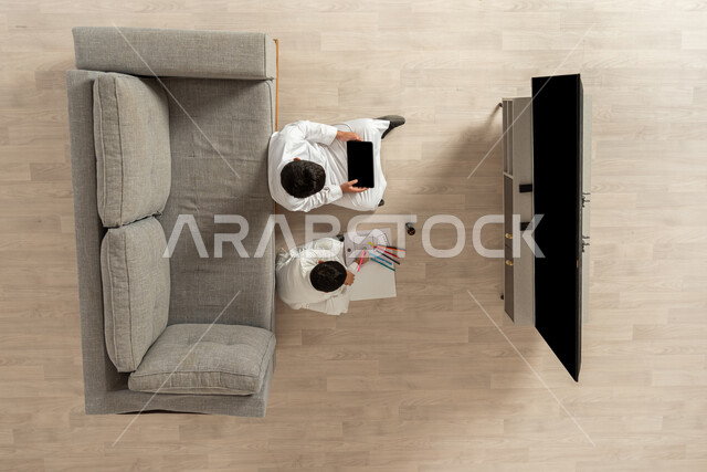 قضاء وقت ممتع في تعلم الرسم والتلوين، النظر الى الكاميرا بإيماءات السعادة والفرح, استغلال أوقات الفراغ بالأنشطة الالكترونية والواقعيه المفيدة، بورتريه علوي عامودي لأخوين عربيين خليجيين سعوديين يجلسان على الأرض في غرفة المعيشة, شاشة سوداء فارغة, خلفية بيج