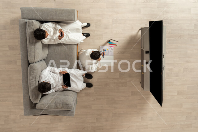 ممارسة الهوايات المسلية, استخدام التقنيات الحديثة في وقت الاستراحة, الترفيه والمتعة في أوقات الفراغ, بورتريه علوي عامودي لمجموعة أولاد عربيون خليجيون سعوديون يرتدون الثوب التقليدي السعودي يجلسون في غرفة المعيشة, الاستمتاع في هواية الرسم والتلوين, خلفية بيج