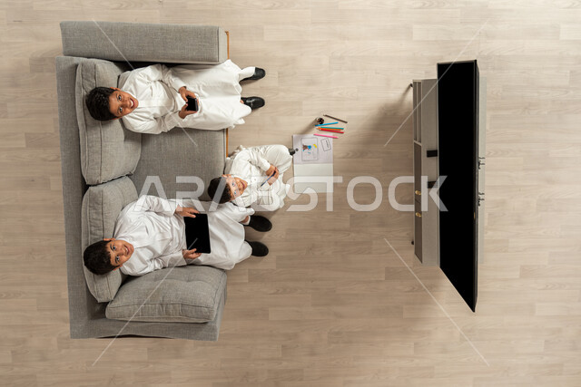 الترفيه والمتعة في أوقات الفراغ, بورتريه علوي عامودي لمجموعة أولاد عربيون خليجيون سعوديون يرتدون الثوب التقليدي  يجلسون في غرفة المعيشة, ممارسة الهوايات المسلية, استخدام التقنيات الحديثة في الاستراحة, النظر الى الكاميرا بإيماءات الفرح والسعادة, خلفية بيج