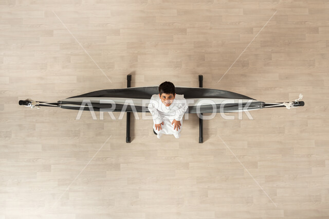 الاسترخاء والمتعة في أوقات الاستراحة، بورتريه علوي عامودي لولد عربي خليجي سعودي يرتدي الثوب التقليدي مستلقي على أرجوحة قماشية معلقة، النظر الى الكاميرا بإيماءات الفرح والسعادة، عطلة صيفية سعيدة, خلفية بيج