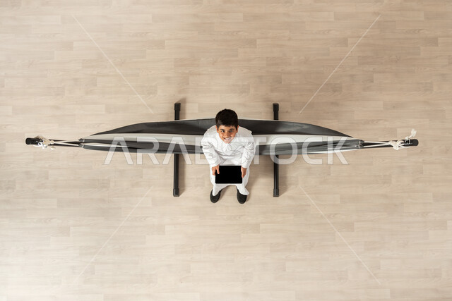 استخدام الأجهزة الحديثة والمتطورة، بورتريه علوي عامودي لولد عربي خليجي سعودي يرتدي الثوب التقليدي مستلقي على أرجوحة قماشية معلقة يحمل بيده الجهاز اللوحي، عرض شاشة سوداء على التابلت، النظر الى الكاميرا بإيماءات السرور والفرح, خلفية بيج