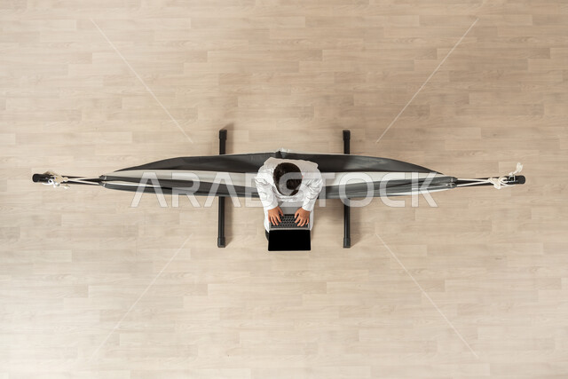 تصفح الانترنت و وسائل التواصل الاجتماعي باستخدام التكنولوجيا، بورتريه علوي عامودي لولد عربي خليجي سعودي يرتدي الثوب التقليدي مستلقي على أرجوحة قماشية معلقة، التواصل و الدردشات عن طريق الحاسوب المحمول، عرض شاشة سوداء فارغة، خلفية بيج