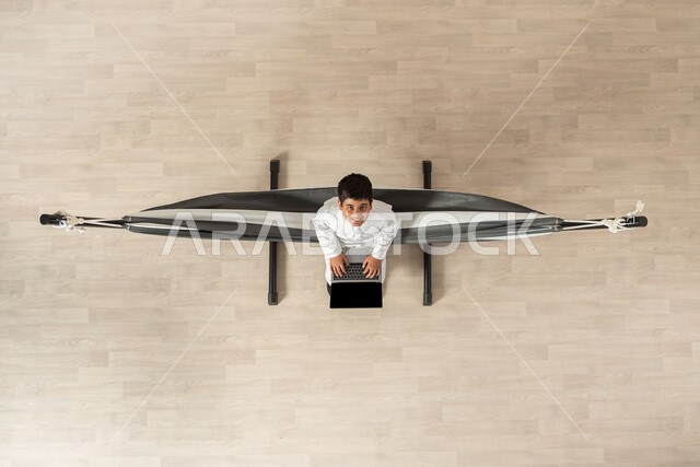 استخدام التقنية والتكنولوجيا الحديثة، بورتريه علوي عامودي لولد عربي خليجي سعودي يرتدي الثوب التقليدي مستلقي على أرجوحة قماشية معلقة, الدراسة عبر الحاسب المحمول، النظر الى الكاميرا بإيماءات السعادة والمتعة، خلفية بيج