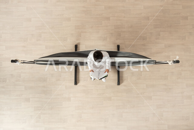 الجمع بين الفائدة والمتعة, ممارسة هواية القراءة، بورتريه علوي عامودي لولد عربي خليجي سعودي يرتدي الثوب التقليدي مستلقي على أرجوحة قماشية معلقة يمسك كتاب بيده، قضاء الوقت بعيداً عن التكنولوجيا, خلفية بيج