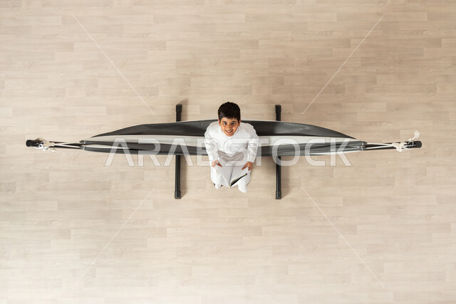 ممارسة المطالعة والقراءة، الجمع بين الفائدة والمتعة, بورتريه علوي عامودي لولد عربي خليجي سعودي يرتدي الثوب التقليدي مستلقي على أرجوحة قماشية معلقة يمسك  بيده كتاب، النظر الى الكاميرا بإيماءات الفرح والسعادة, خلفية بيج