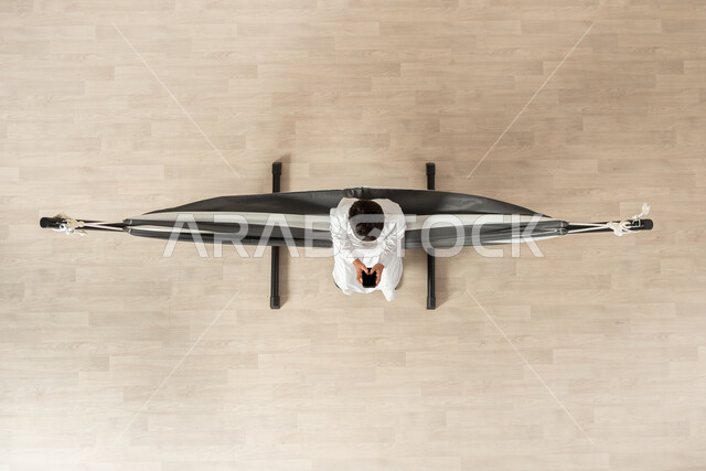 استخدام أحد وسائل التواصل، بورتريه علوي عامودي لولد عربي خليجي سعودي يرتدي الثوب التقليدي مستلقي على أرجوحة قماشية معلقة يمسك هاتفه المحمول بيده، عرض شاشة سوداء فارغة عبر الجوال، التواصل مع الأهل والأصدقاء, تبادل الأحاديث والأخبار الجيدة, خلفية بيج