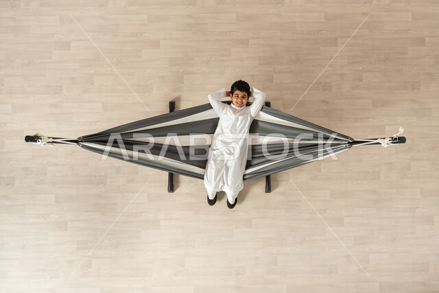 قضاء أوقات ممتعة في الاستراحة، بورتريه علوي عامودي لولد عربي سعودي خليجي يرتدي الثوب التقليدي يستلقي على أرجوحة قماشية معلقة و ينظر الى الكاميرا، يديه خلف رأسه ضاحك الوجه، خلفية بيج