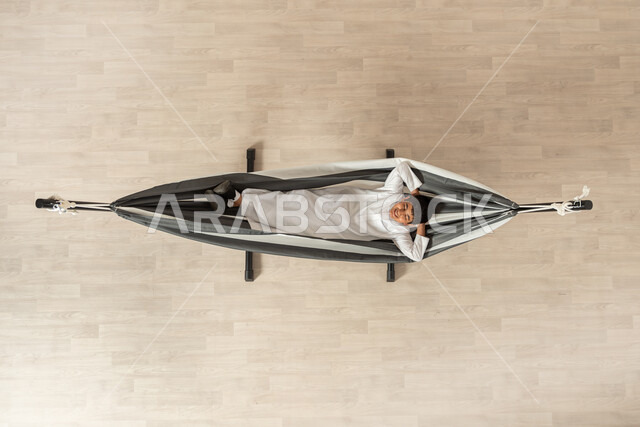 الحاجة إلى الراحة و الهدوء، بورتريه علوي عامودي لولد عربي سعودي خليجي يرتدي الثوب التقليدي مستلقي على أرجوحة قماشية معلقة، يديه خلف رأسه وإيماءاته تدل على الفرح والسعادة، خلفية بيج