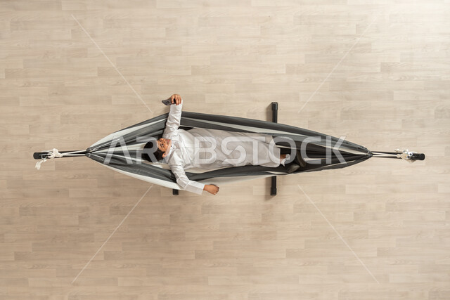 المتعة في الاستلقاء والراحة، بورتريه علوي عامودي لولد عربي سعودي خليجي يرتدي الثوب التقليدي يستلقي على أرجوحة قماشية معلقة، التقاط الصور الفوتوغرافية من خلال الهاتف المحمول، خلفية بيج