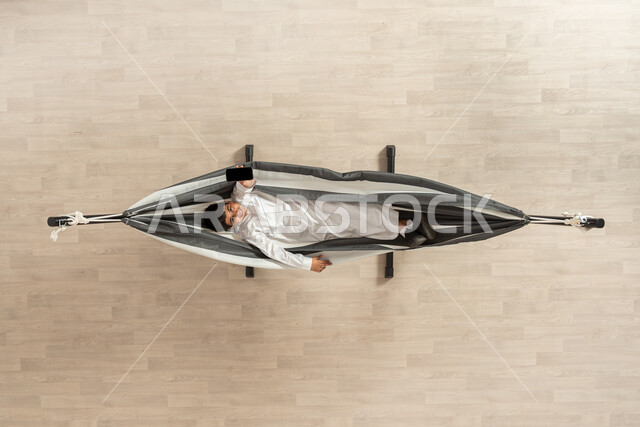 ممارسة الهوايات في اوقات الفراغ، بورتريه علوي عامودي لولد عربي سعودي خليجي يرتدي الثوب التقليدي يستلقي على أرجوحة قماشية معلقة ينظر الى الكاميرا، الابتسامة على وجهه و يحمل هاتف محمول موجه للأعلى، خلفية بيج