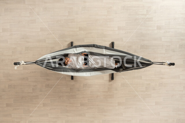 الاعتماد على التكنولوجيا والأجهزة الحديثة، التواصل مع الأصدقاء، بورتريه علوي عامودي لرجل عربي خليجي سعودي يرتدي الثوب السعودي التقليدي يسلتقي على أرجوحة قماشية معلقة، يحمل بيديه جهاز التابلت وينظر للكاميرا، الاستمتاع بوقت الفراغ، ايماءات السعادة والرضا، خلفية بيج