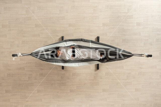 استخدام جهاز تقني، تصفح الإنترنت بسهولة، بورتريه علوي عامودي لشاب عربي خليجي سعودي يستمتع  باستخدام( التابلت) يرتدي اللباس السعودي التقليدي يجلس على سرير قماشي معلق، التسويق عن بعد، خلفية بيج