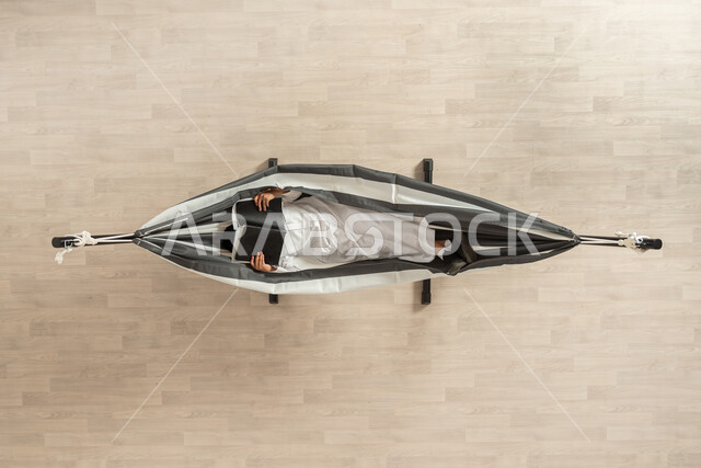 التمتع بالاسترخاء بعد المطالعة،  بورتريه عامودي علوي لشاب عربي خليجي سعودي يرتدي زي سعودي تقليدي يستلقي على أرجوحة قماشية معلقة، يضع الكتاب على وجهه، الاستمتاع بالاسترخاء بعد القراءة، قراءة القصص والروايات الممتعة، خلفية بيج