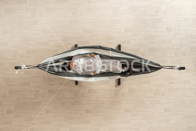 The hobby of reading and educating oneself, spending free time entertaining oneself through reading, an overhead vertical portrait of a Saudi Gulf Arab man wearing the traditional Saudi dress sitting on a hanging cloth hammock, holding a book and enjoying reading, beige background