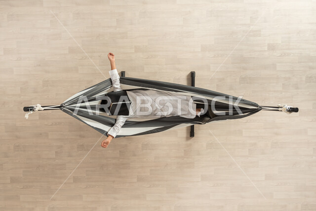 الاستمتاع بوقت من الراحة بعد المطالعة، بورتريه عامودي علوي لشاب  عربي خليجي سعودي يرتدي اللباس السعودي التقليدي يستلقي على سرير قماشي معلق، يضع الكتاب على وجهه ويمد يديه، الاستمتاع بالجلوس مع الكتاب، خلفية بيج