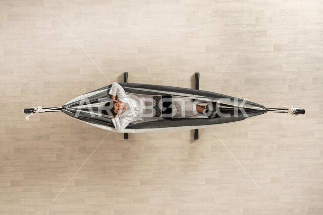 قضاء الوقت بالاسترخاء و السكون بورتريه عامودي علوي لشاب عربي خليجي سعودي يرتدي الثوب التقليدي،  و يقوم بالاستلقاء على أرجوحة قماشية معلقة وينظر للكاميرا، استخدام التقنيات الحديثة، العمل عن بعد، خلفية بيج