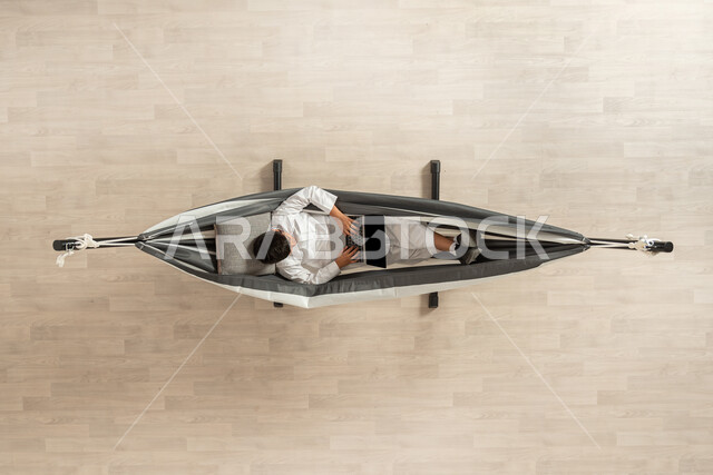 العمل على الحاسوب المحمول،بورتريه علوي عامودي لشاب عربي خليجي سعودي يرتدي الثوب السعودي التقليدي،  يسلتقي على الأرجوحة القماشية المعلقة ويستخدم اللاب توب، استخدام الأجهزة الحديثة، خلفية بيج