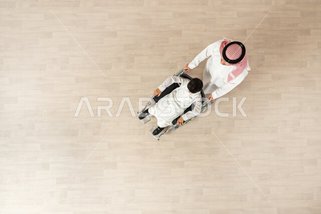 اهتمام ودعم المجتمع السعودي لذوي الاحتياجات الخاصة، تقديم الحب والرعاية لذوي الهمم، مساعدة مرضى الإعاقة الحركية على التنقل بسهولة, بورتريه  علوي عامودي لولد سعودي مقعد يجلس على كرسي متحرك بمساعدة والده، العطف والحب بين أفراد الأسرة، خلفية بيج