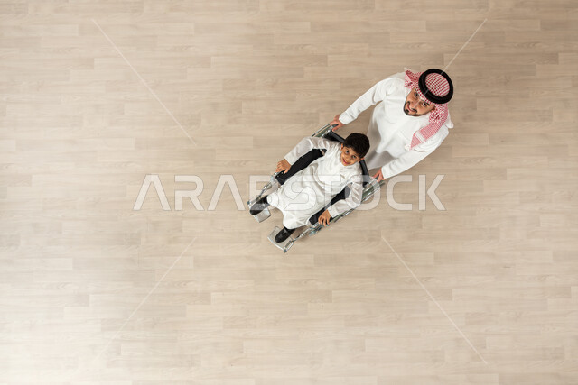 مساعدة مرضى ذوي الإعاقة الحركية على التنقل بسهولة, شعور ذوي الهمم بالراحه والطمأنينه في بيئتهم, بورتريه علوي عامودي لولد سعودي خليجي يرتدي الثوب التقليدي يجلس على كرسي متحرك برفقة والده، العطف والحب بين أفراد الأسرة، الاهتمام بالجانب النفسي والجسدي للمقعدين، خلفية بيج