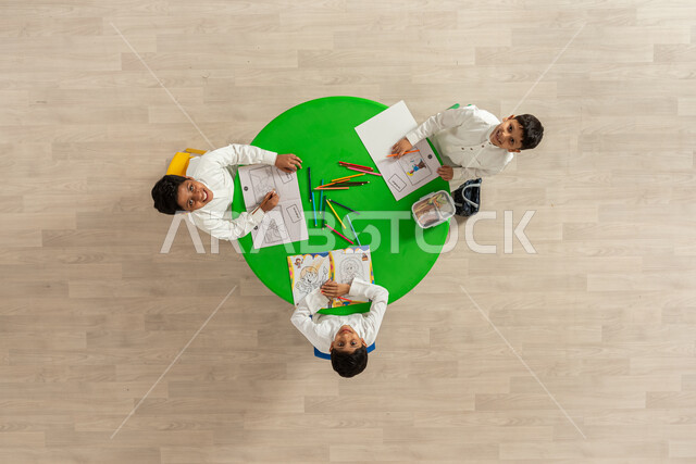 Entertaining entertainment activities, vertical portrait of students from the Kingdom of Saudi Arabia, wearing traditional Saudi clothing, drawing and coloring and looking at the camera, the concept of interest in hobbies, facial gestures indicating happiness, beige background