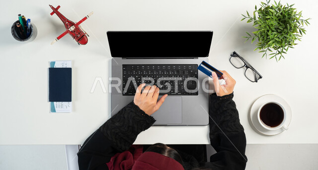 الولوج الى المستلزمات اللازمة عن طريق الاونلاين، السياحة والقيام بالجولات الترفيهية، الاستعداد لبدء الرحلة في العطلة الصيفية، بورتريه علوي عامودي لإمرأة عربية خليجية سعودية تستخدم الحاسوب بجانبها جواز وتأشيرة السفر، الدفع عبر بطاقة المصرف البنكية، خلفية بيضاء