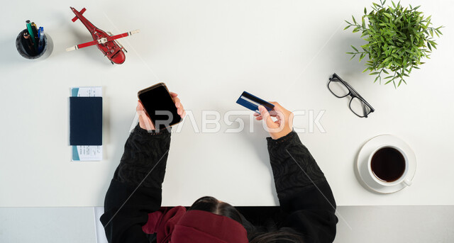 Preparing to start the trip during the summer vacation, tourism and recreational tours, a vertical overhead portrait of a Saudi Gulf Arab woman using the phone next to her passport and travel visa, paying via bank card, obtaining the necessary supplies via the Internet, white background.