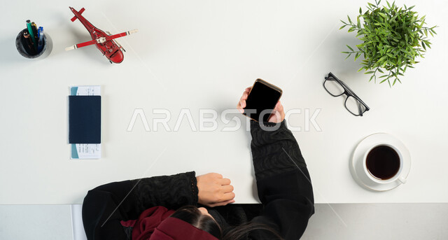 The concept of tourism and taking recreational tours, preparing for an enjoyable summer trip, a vertical overhead portrait of a Saudi Gulf Arab woman using the phone with her passport and travel visa next to her, obtaining the necessary supplies via the Internet, integrating technology into daily life, white background.