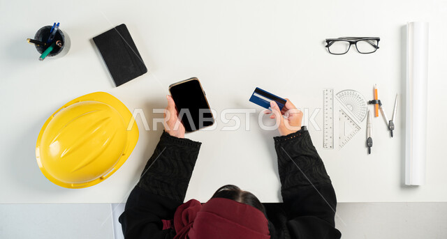 الحصول على المستلزمات اللازمة عن طريق الاونلاين، مفهوم الهندسة والإنشاءات المعمارية، التسوق عبر المواقع الالكترونية، بورتريه علوي عامودي لإمرأة عربية خليجية سعودية تستخدم الهاتف بجانبها خوذة الحماية والأدوات الهندسية، الدفع عبر بطاقة المصرف البنكية، خلفية بيضاء